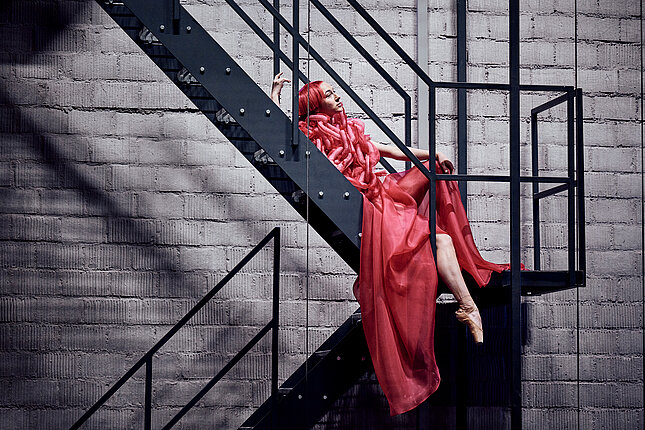 Tanečnice s červenými vlasy a v červených šatech pózuje na ocelovém schodišti.
